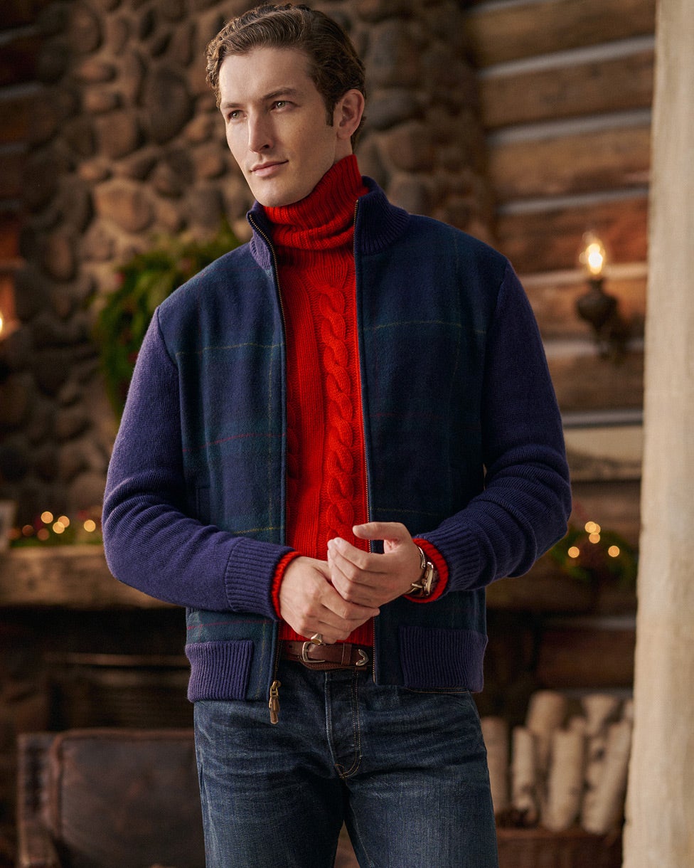 Man in een rode kabeltrui met turtleneck, een marineblauw jack en een spijkerbroek.