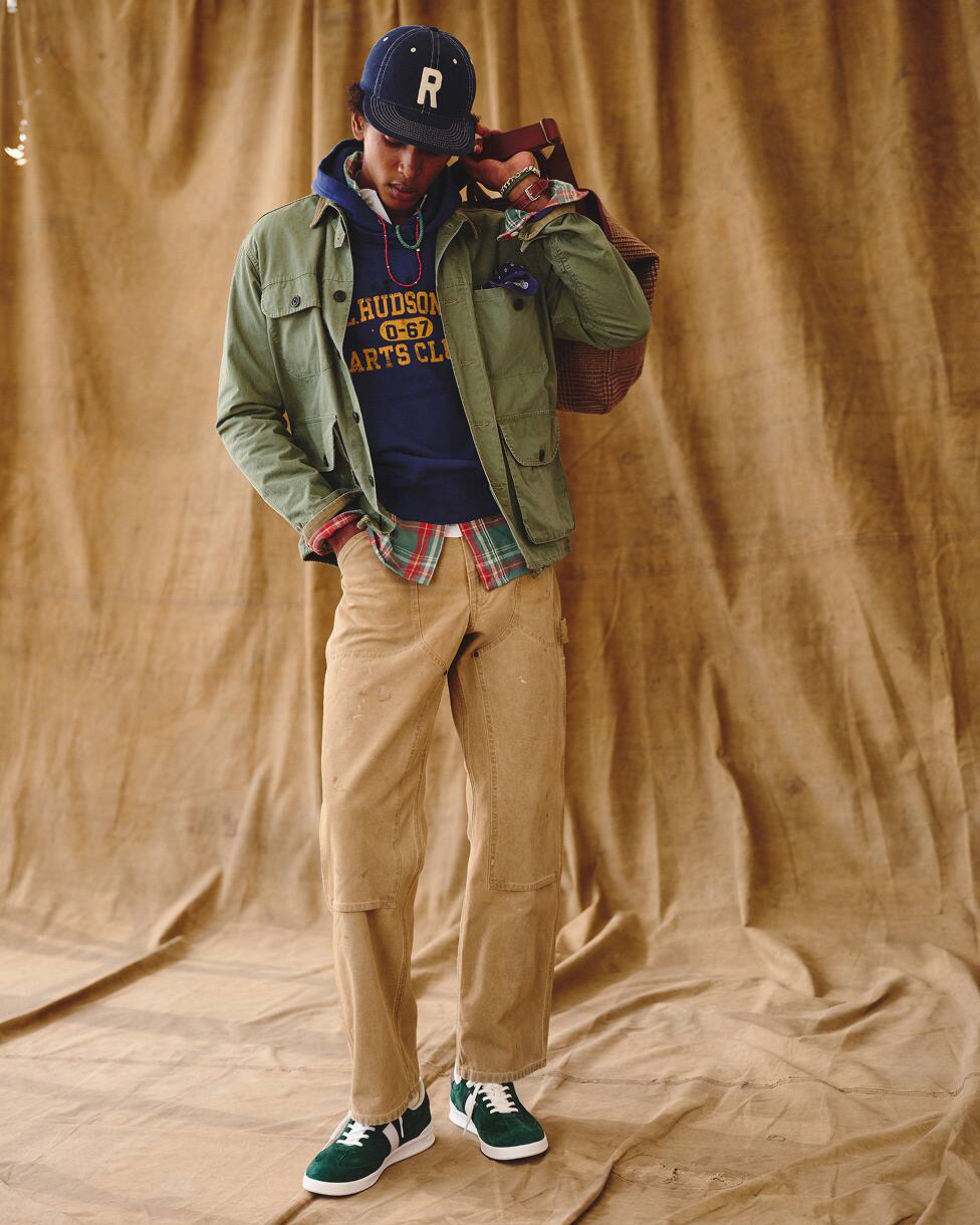 Een man in een klusjas over een hoodie en een geruit overhemd, met een beige broek, sneakers en een pet.
