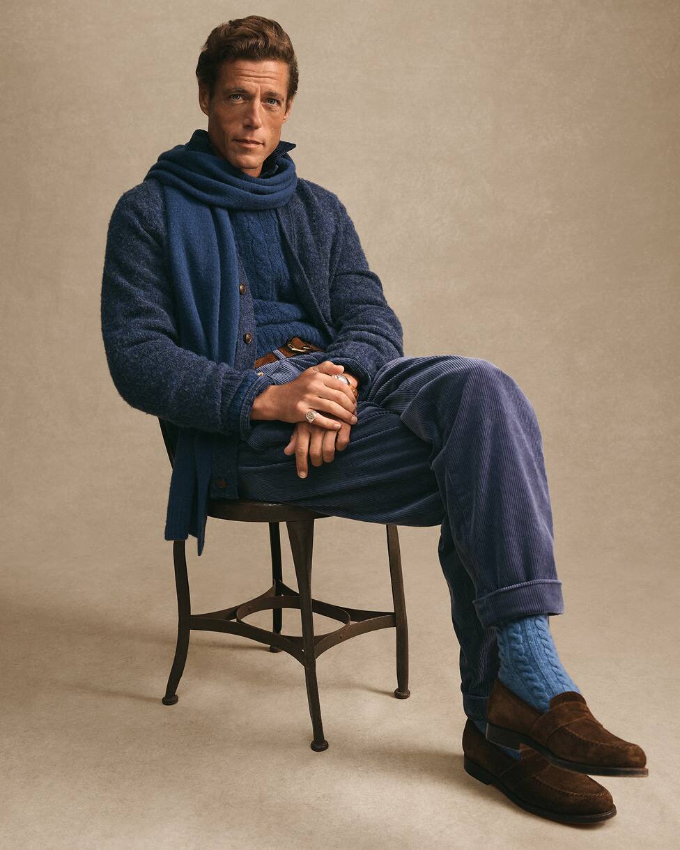 Zittende man gekleed in een blauwe trui en bijpassende sjaal, met bruine loafers.