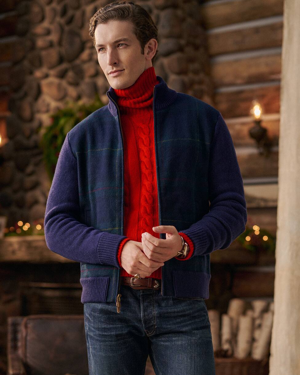 Man in een rode kabeltrui met turtleneck, een marineblauw jack en een spijkerbroek.